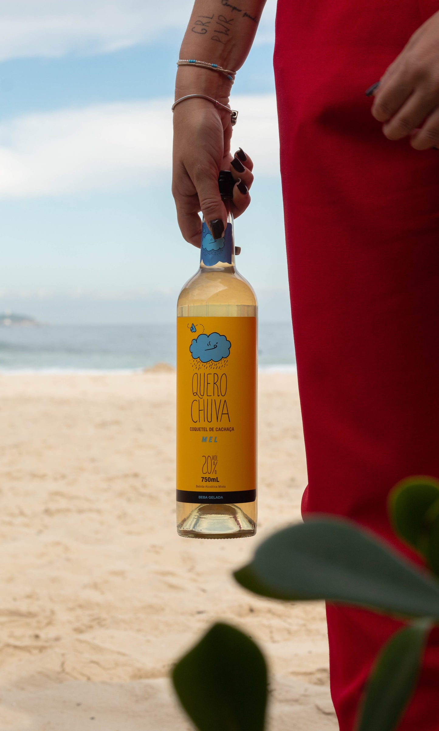 Quero Chuva Mel 750 ml na praia — garrafa na mão, clima de verão