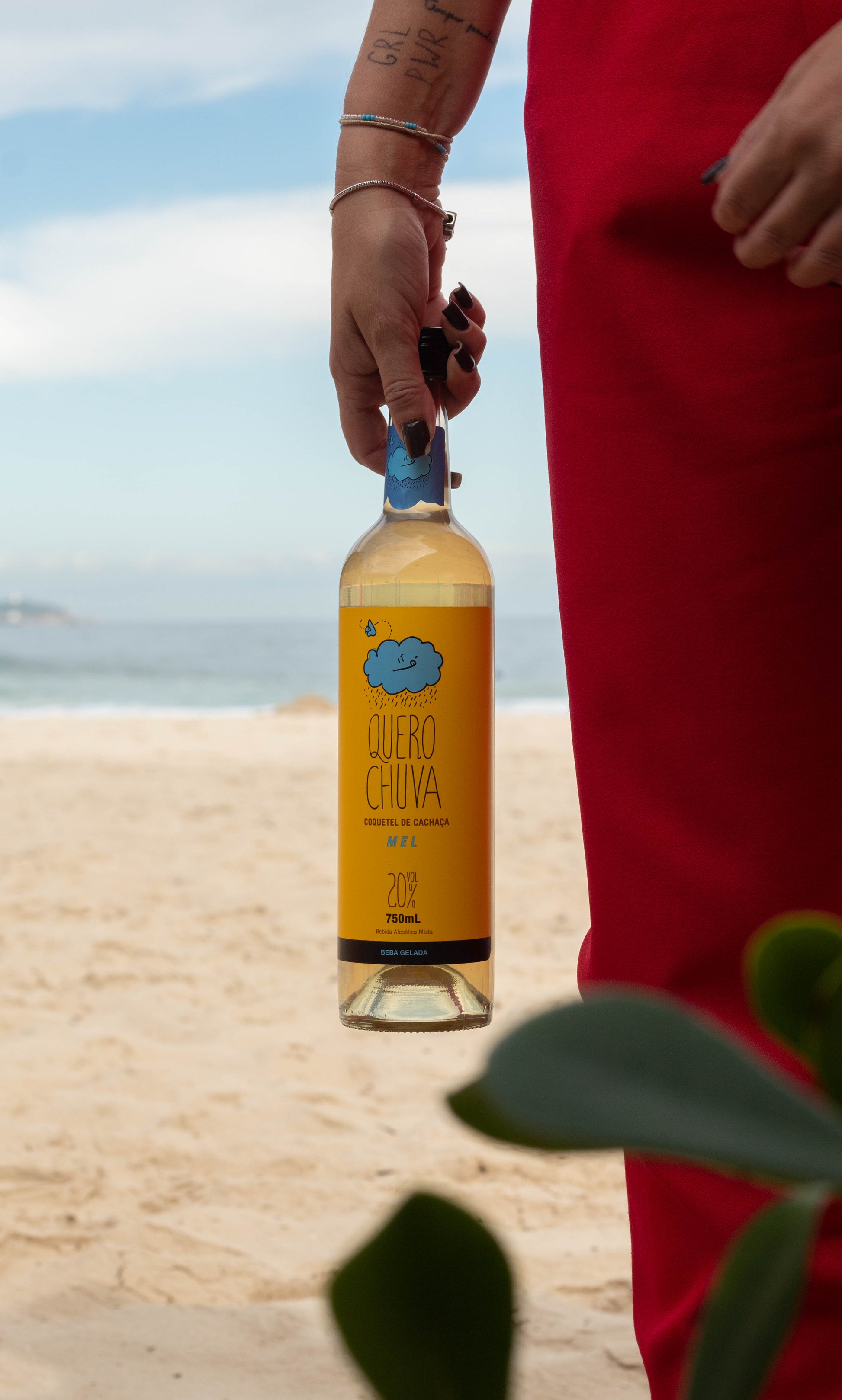 Quero Chuva Mel 750 ml na praia — garrafa na mão, clima de verão